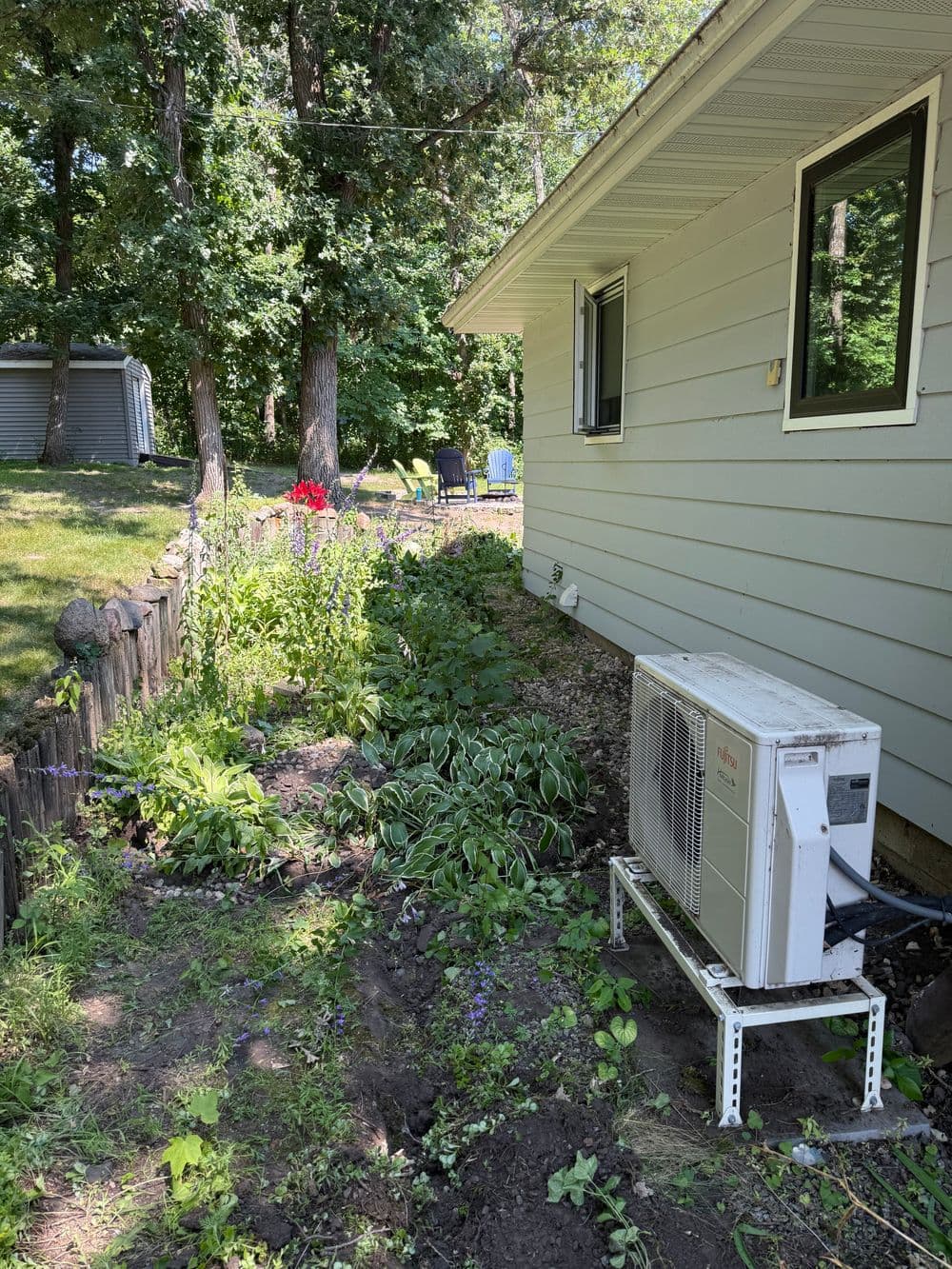 Residential garden with flowering plants and an air conditioning unit beside a house.