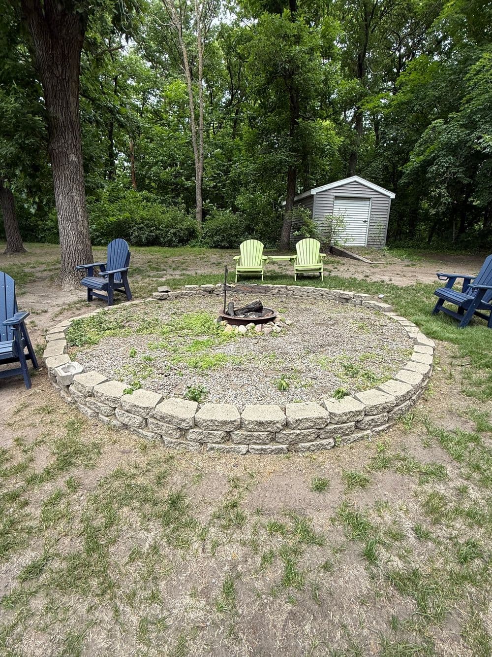 Fire pit surrounded by circular stone seating area with colorful chairs in a forested setting.